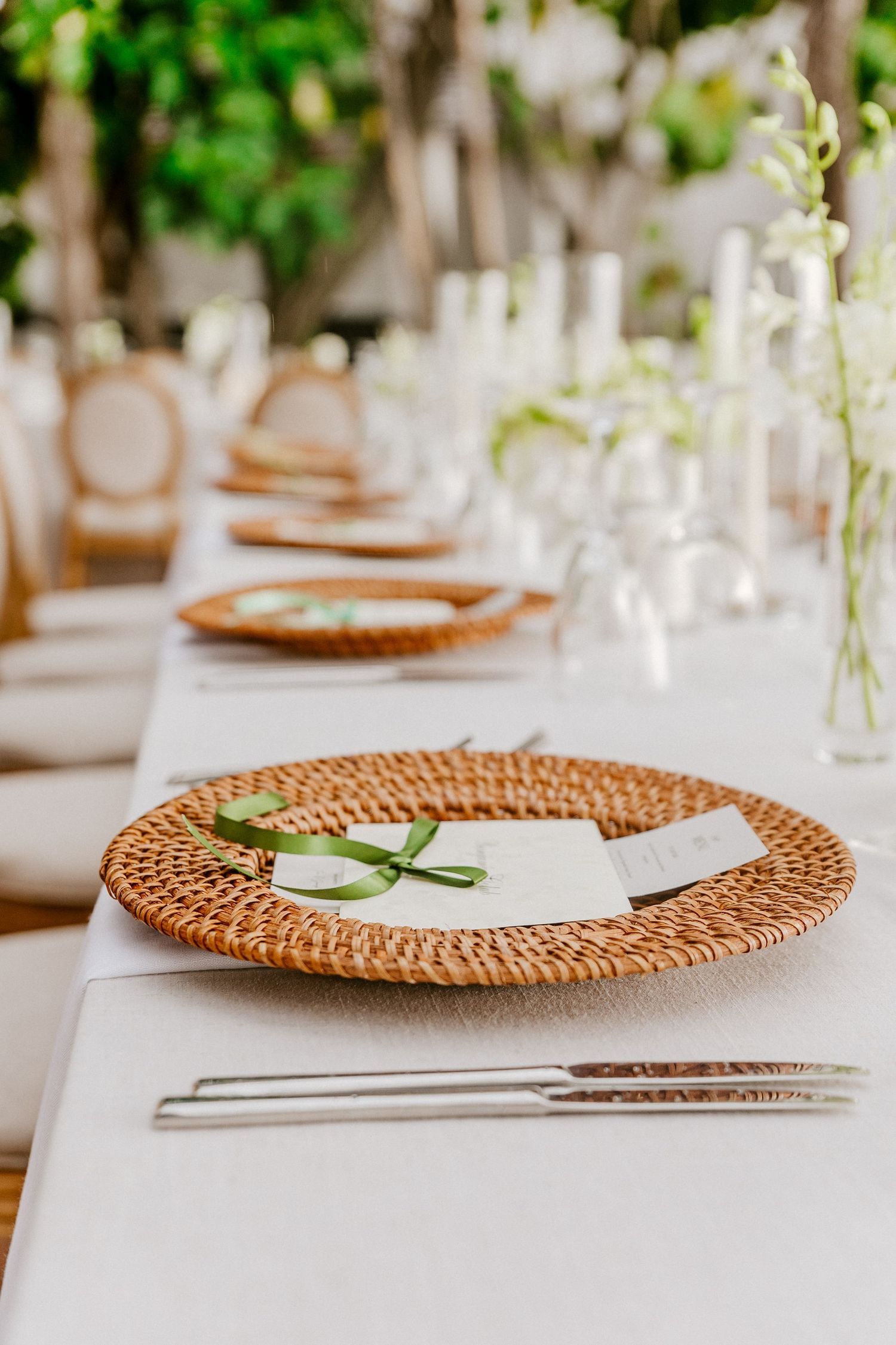 Long outdoor wedding reception table with wicker charger plates, white menus tied with green ribbon, and floral centerpieces.