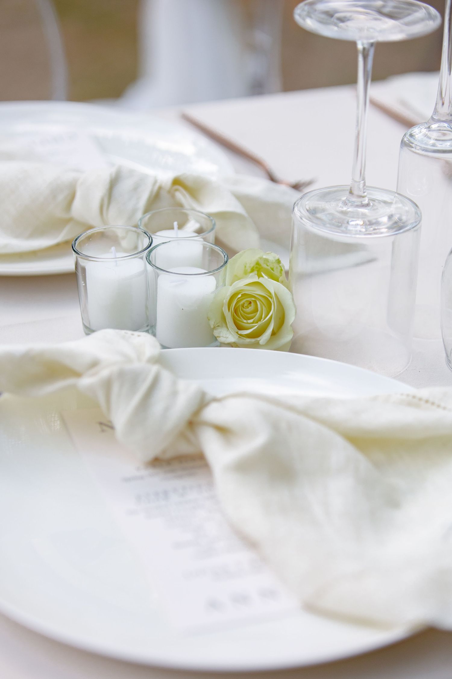 Elegant white wedding table setting with cream linen napkin, white rose, votive candles, and crystal wine glasses on white plate.