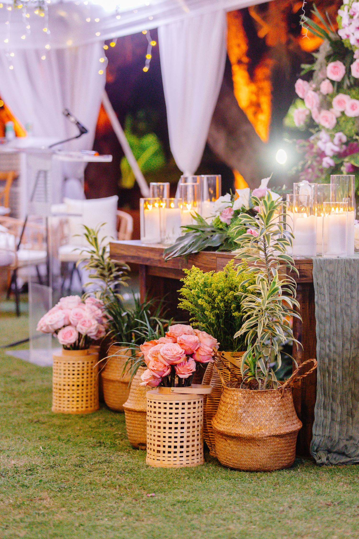 Elegant outdoor wedding decor with pink peonies in wicker baskets, candles, and string lights under a draped tent.