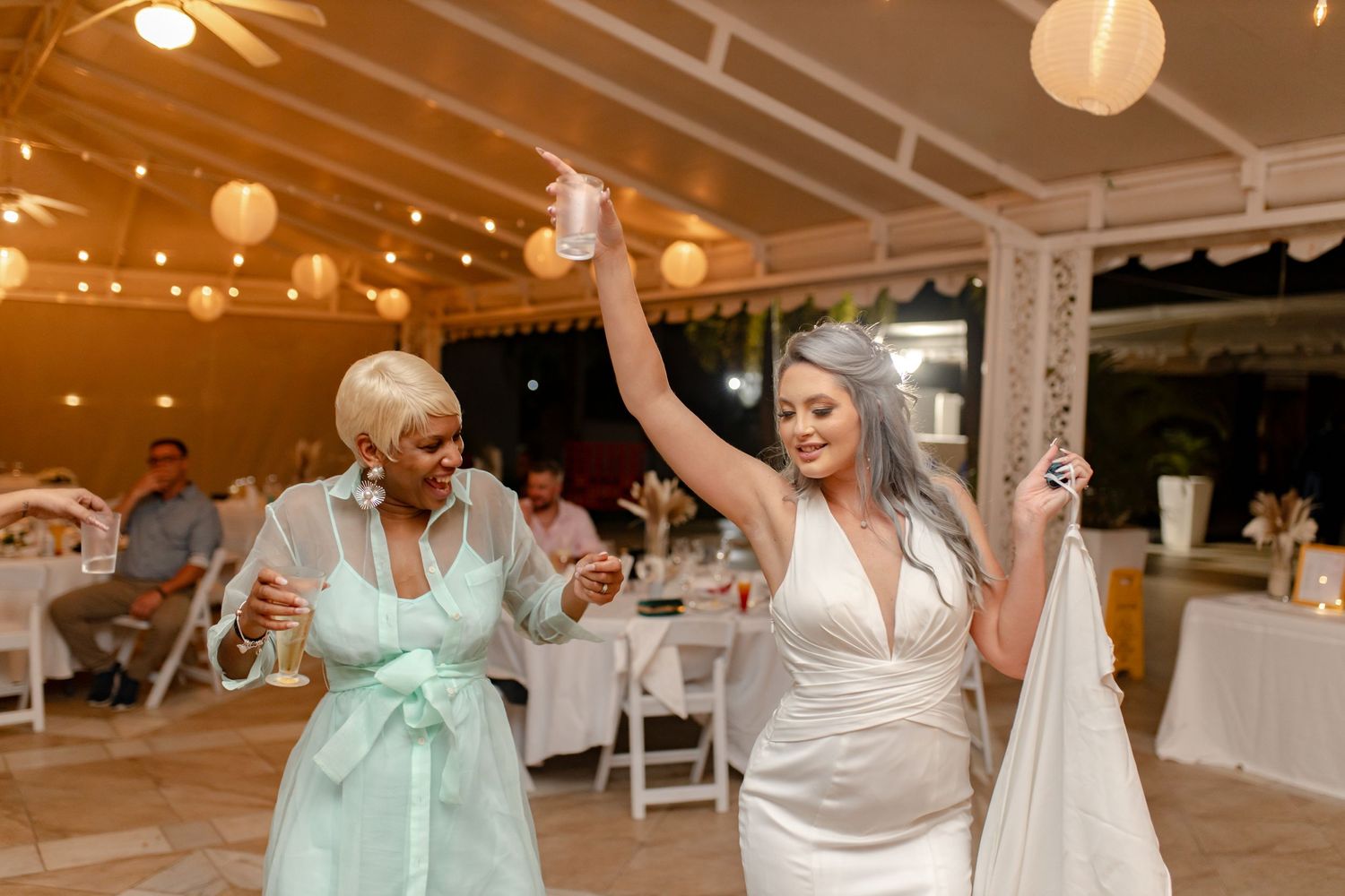 Bride in white gown dances joyfully with guest in mint dress at elegant wedding reception.