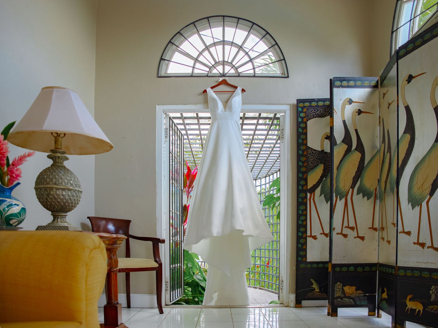 Elegant white wedding dress hanging in sunlit doorway of tropical villa with decorative Asian screen.