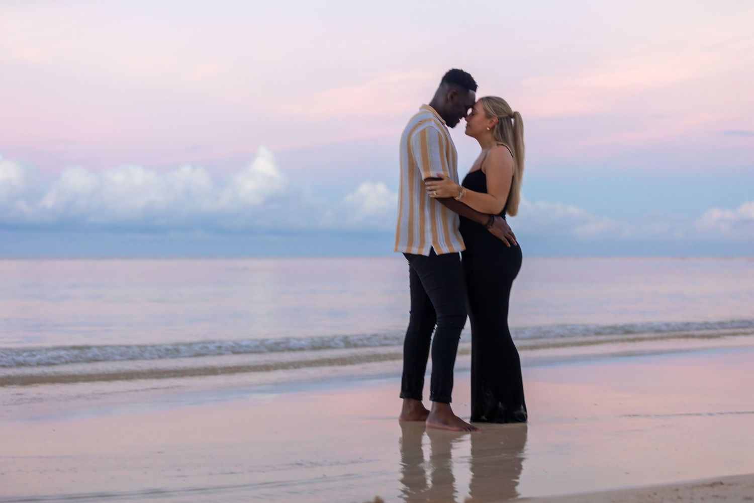 Couple sharing an intimate moment on a sandy beach with a beautiful pastel sunset backdrop.
