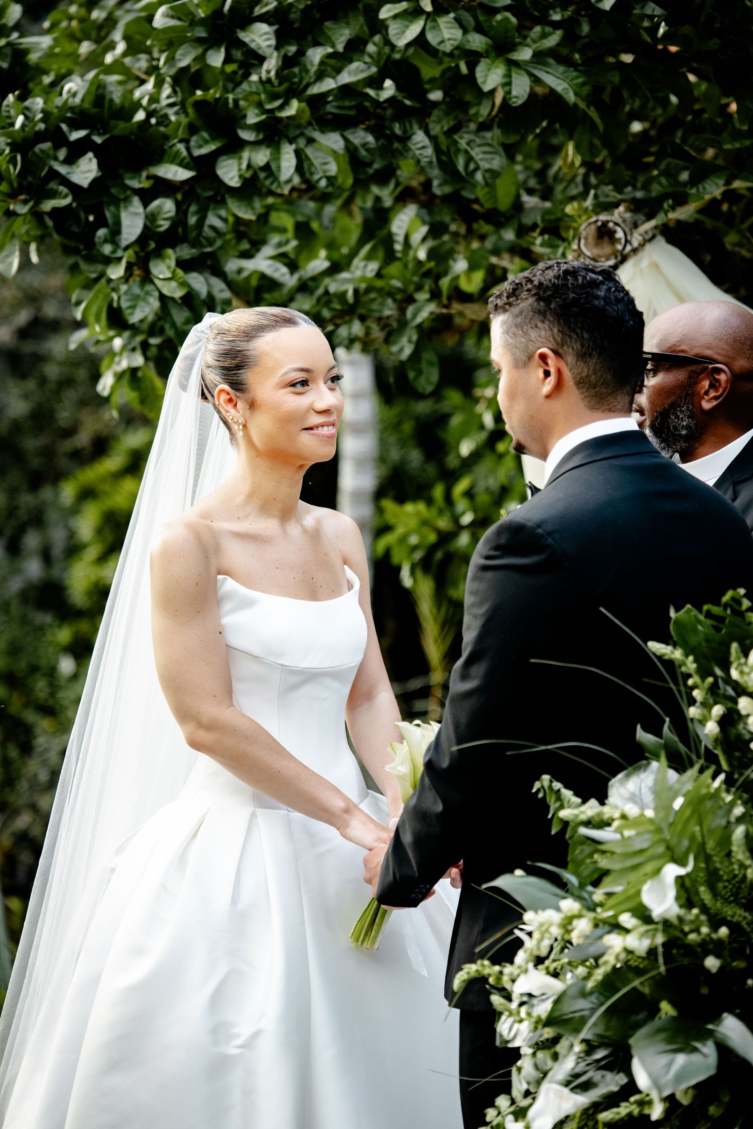 Radiant bride in white strapless gown and veil holding hands with groom during outdoor ceremony.