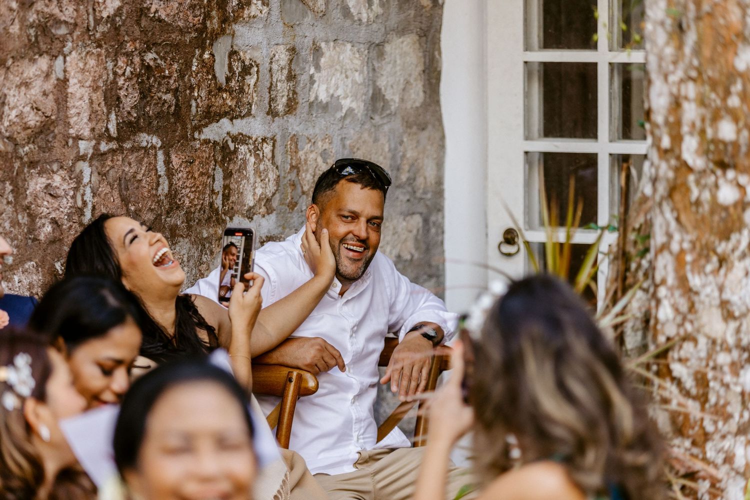 Guests laughing and enjoying a casual outdoor wedding reception against a rustic stone wall.