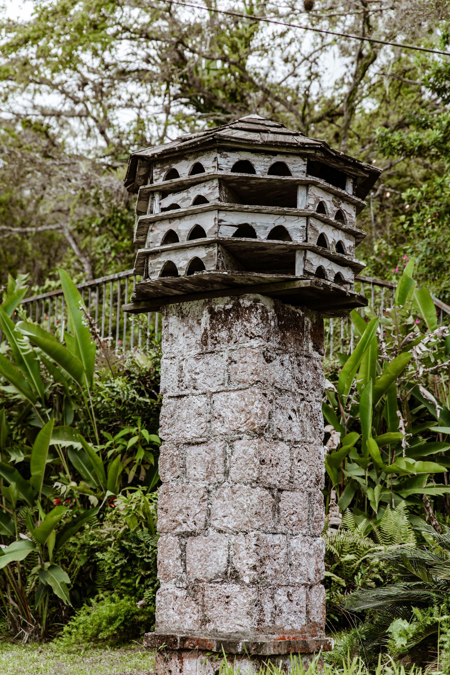 Ornate stone birdhouse on tall brick pillar surrounded by tropical green foliage in garden setting.