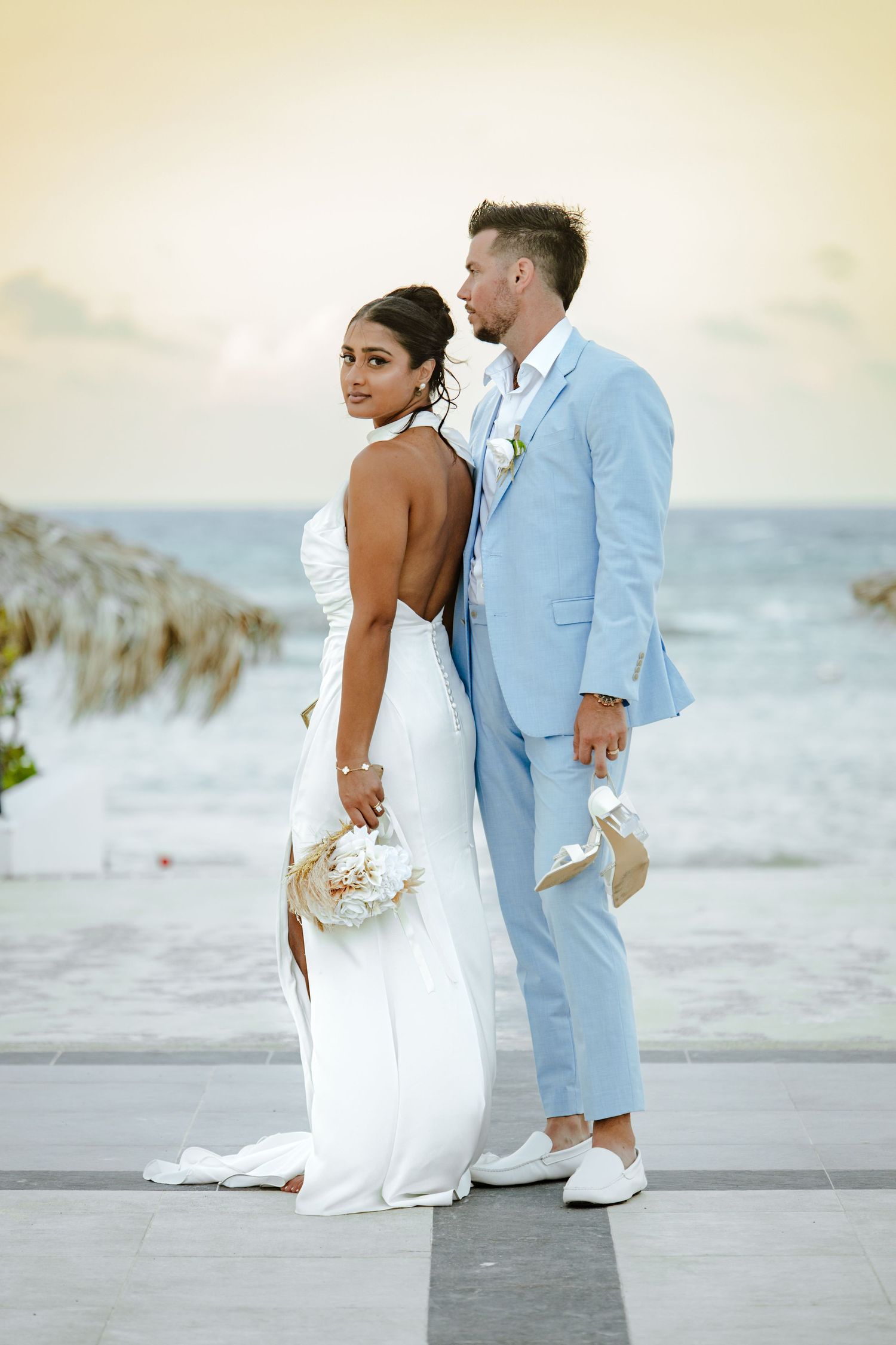 Bride in backless white gown and groom in light blue suit pose together at a beachside wedding venue.