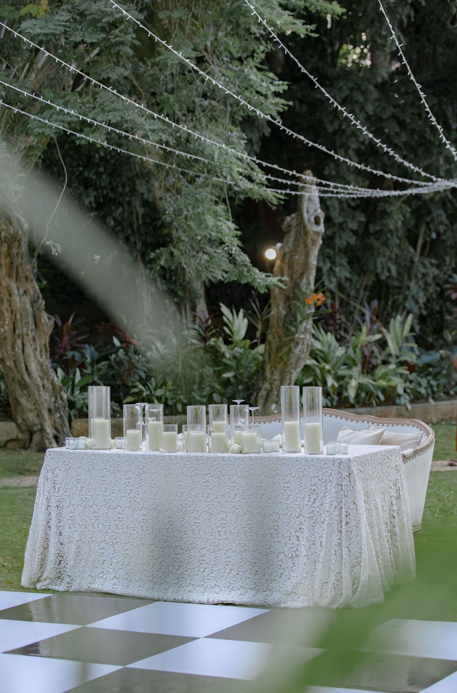 Elegant outdoor wedding reception table with white lace cloth, cylinder candles, and fairy lights strung above a checkered dance floor.
