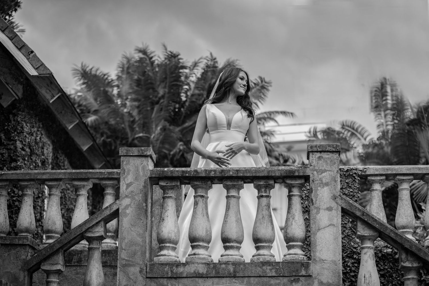 Black and white portrait of bride in wedding gown standing gracefully on decorative stone balustrade.