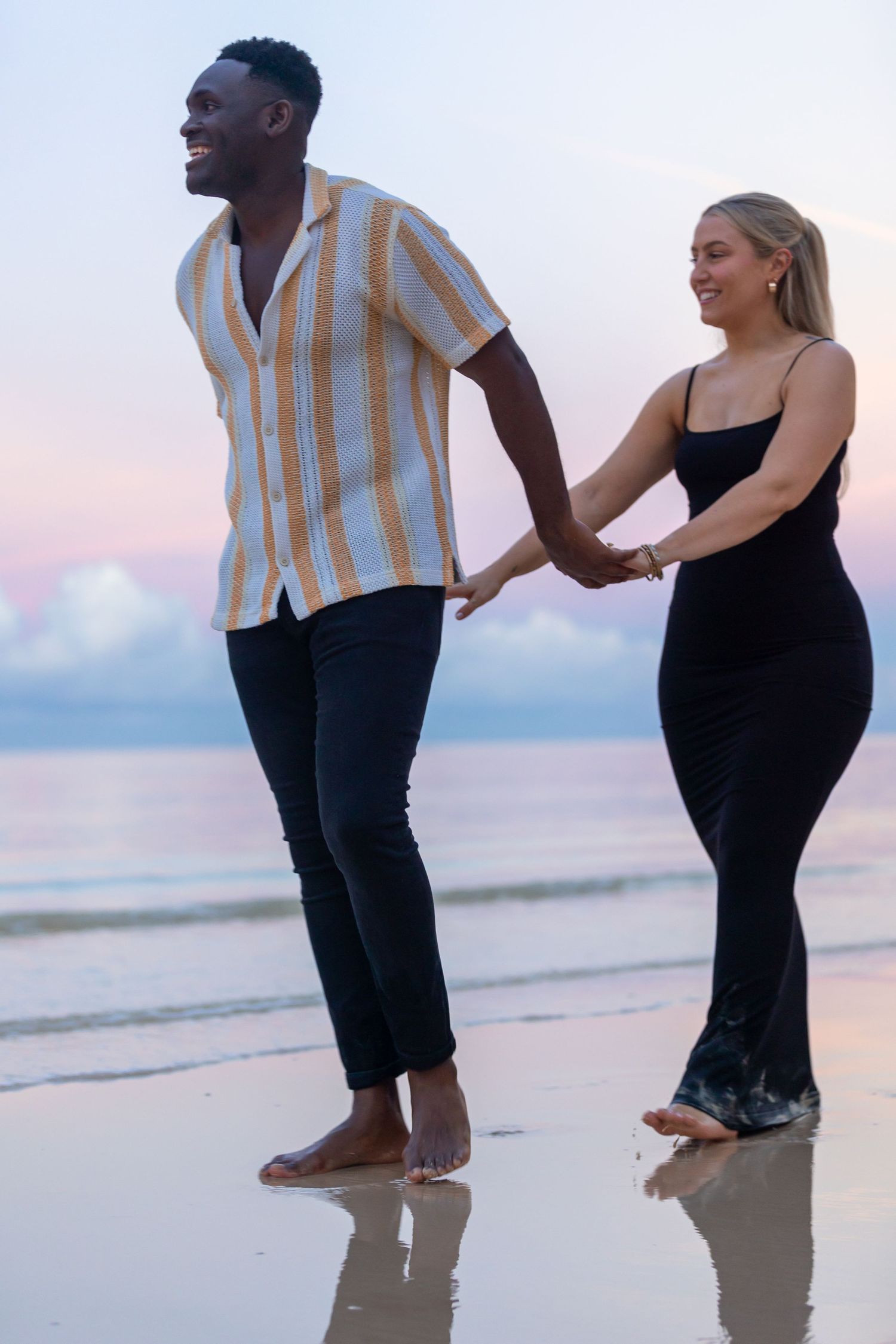 Happy interracial couple holding hands and walking on a scenic beach at pastel sunset.