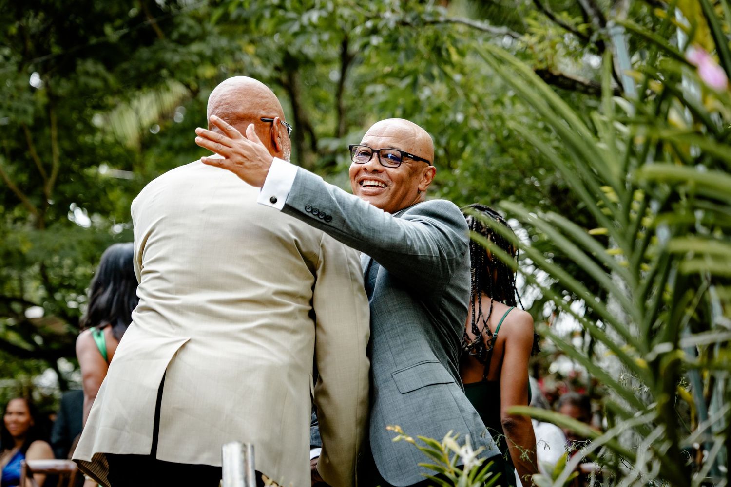 Two men sharing a warm embrace during an outdoor wedding ceremony surrounded by lush greenery.