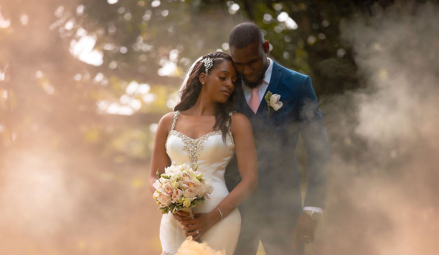Black couple embracing in dreamy smoke-filled garden, bride in lace gown holding pink bouquet, groom in blue suit.