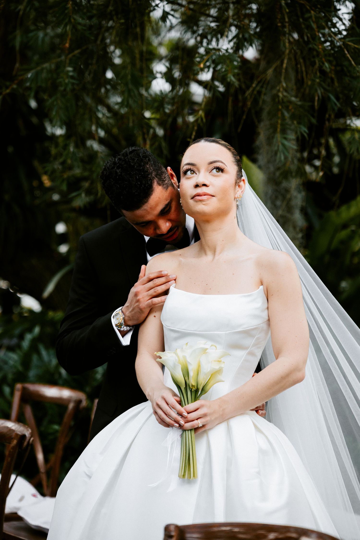 Bride in strapless white ball gown holding calla lily bouquet with groom whispering in her ear.