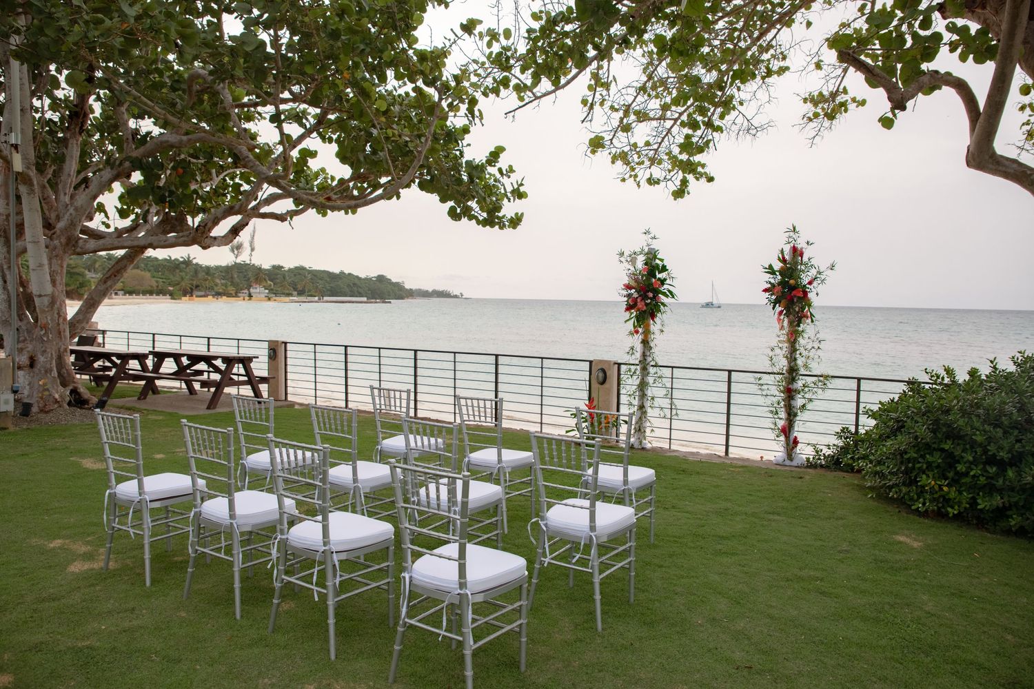 Intimate beachside wedding ceremony with silver chiavari chairs arranged under large tree overlooking the ocean.