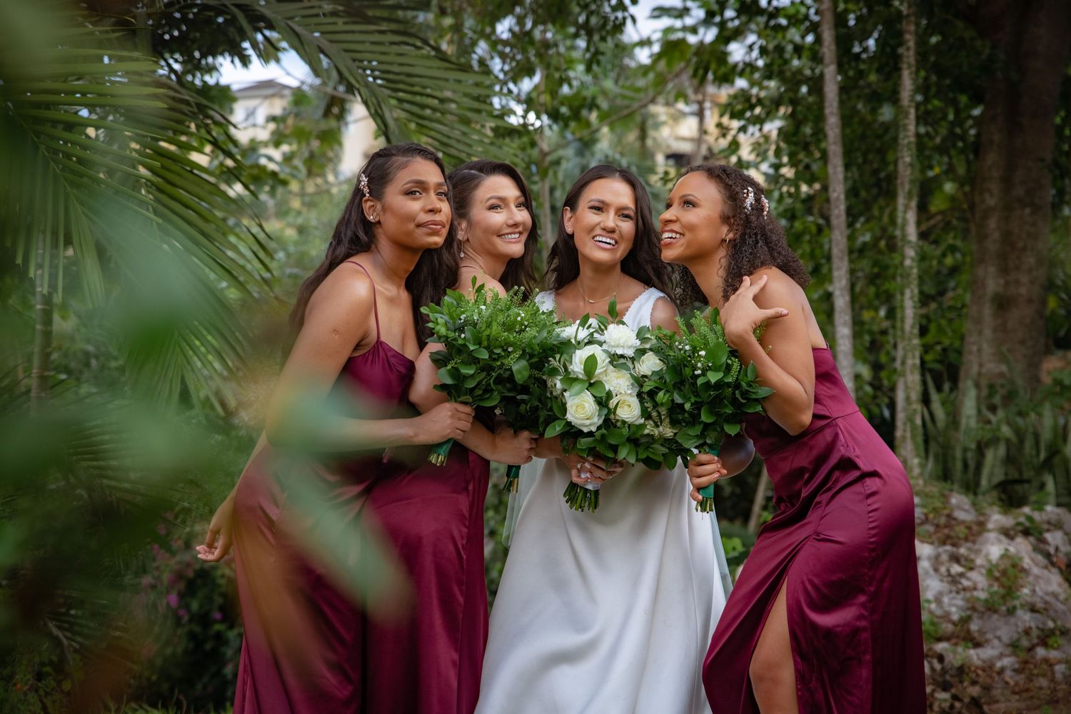 Smiling bride holding white bouquet surrounded by three bridesmaids in wine-colored dresses in garden.