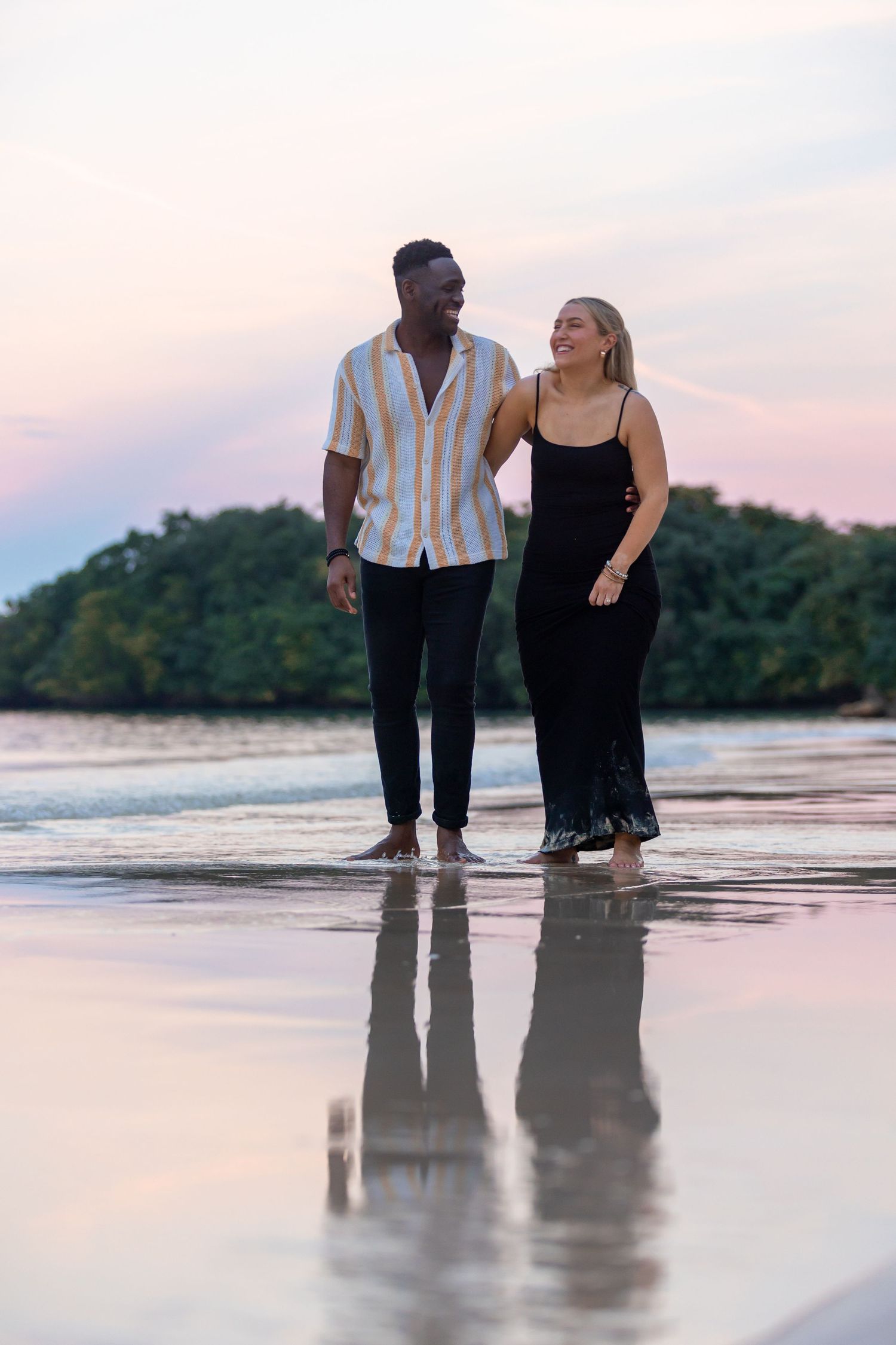 Smiling interracial couple walking arm in arm on a reflective beach at pastel sunset.