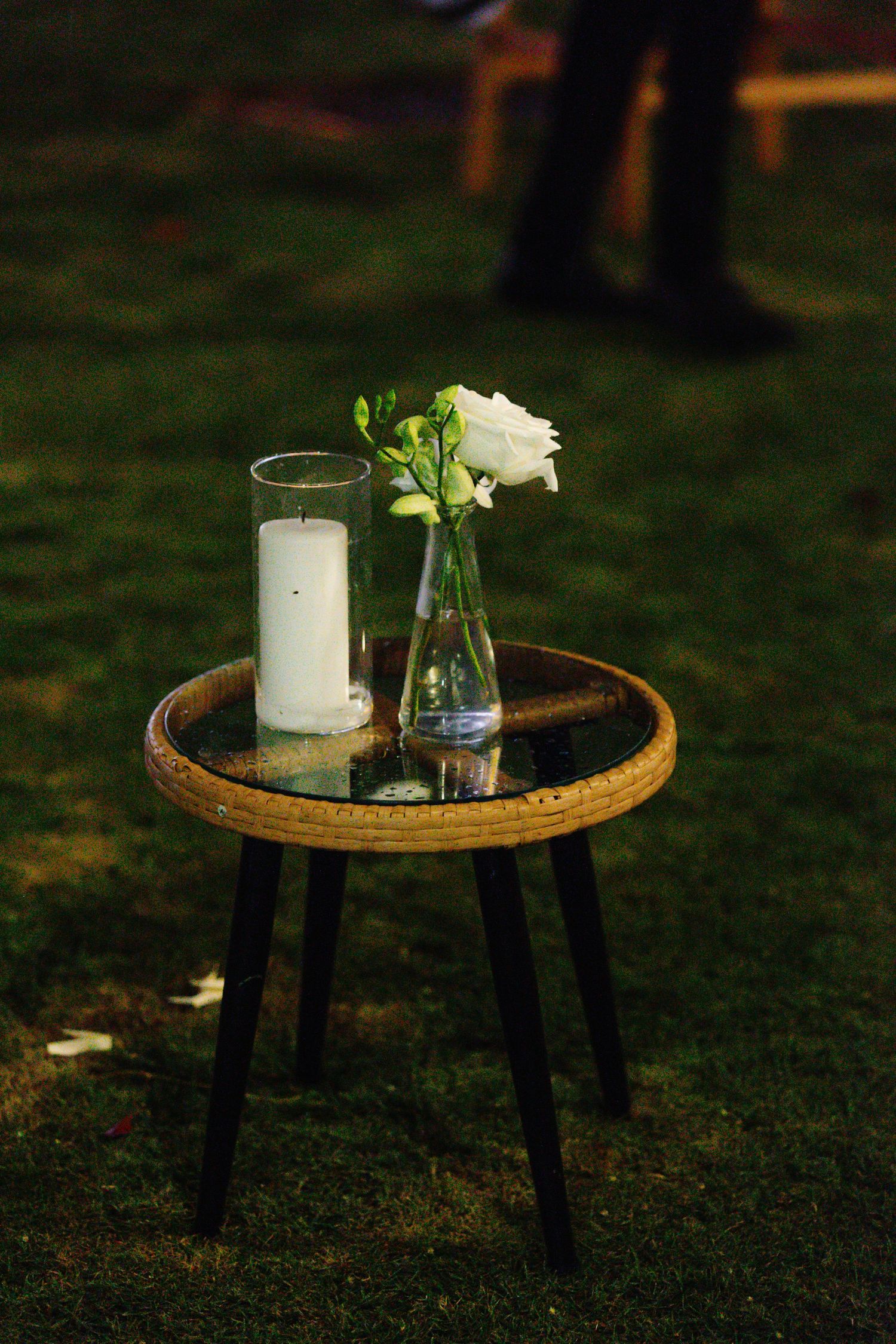 Small rattan side table with a white pillar candle and single white rose in a glass vase on grass.