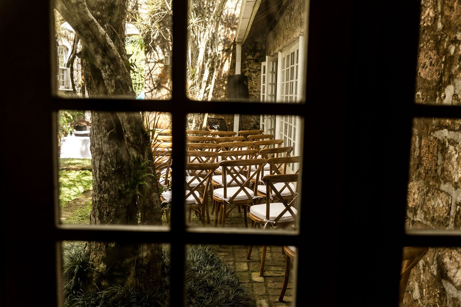 View through a dark window frame of a rustic outdoor patio with wooden chairs surrounded by large trees.