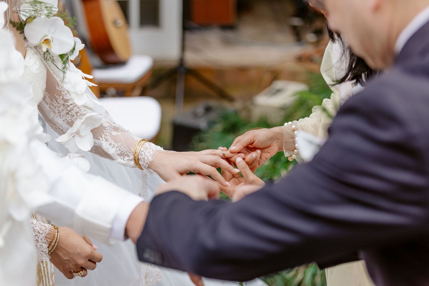 Groom placing a wedding ring on bride's finger during a ceremony, bride wearing lace dress with floral accessories.