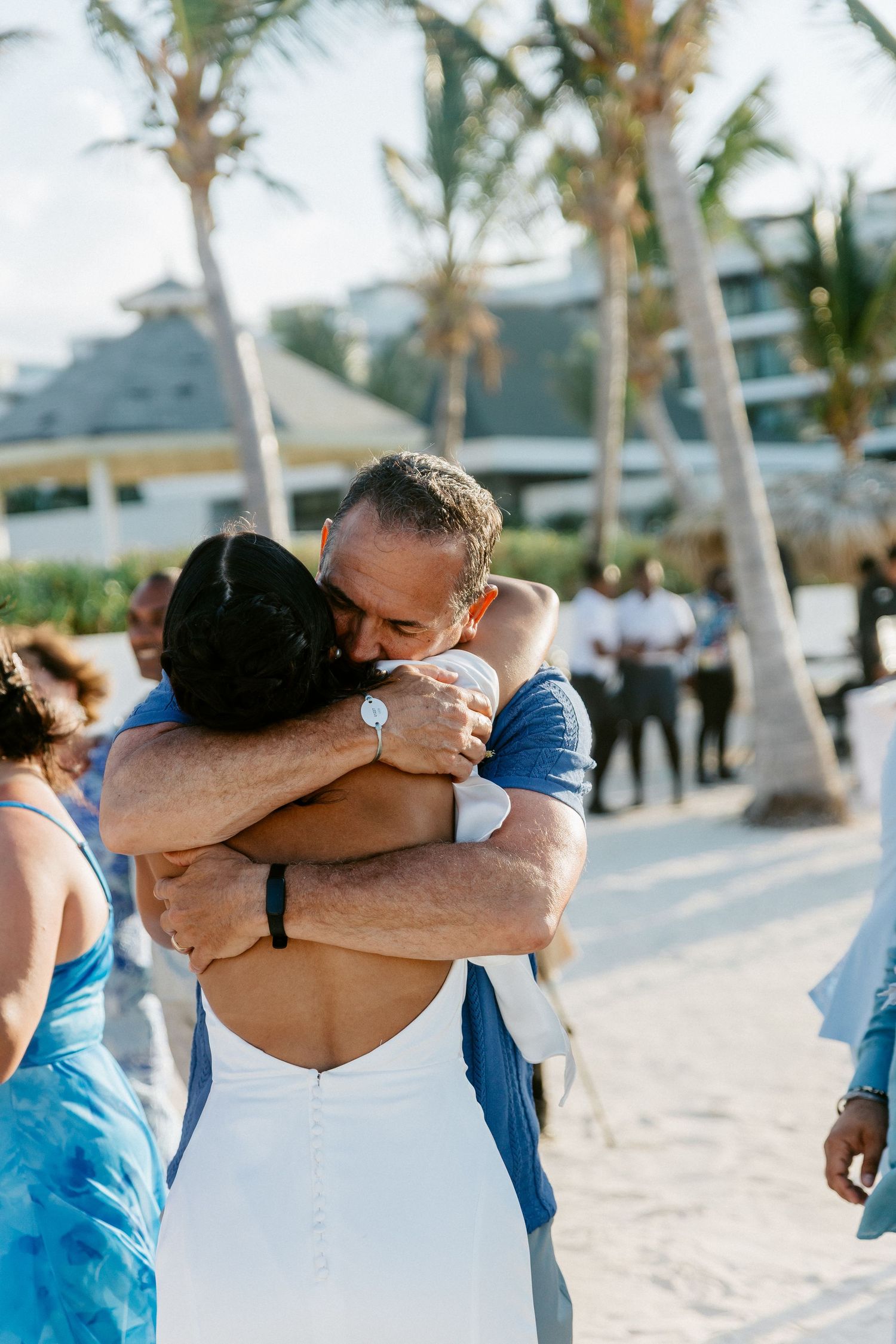 Emotional wedding moment as bride in white backless gown embraces guest on tropical beach.