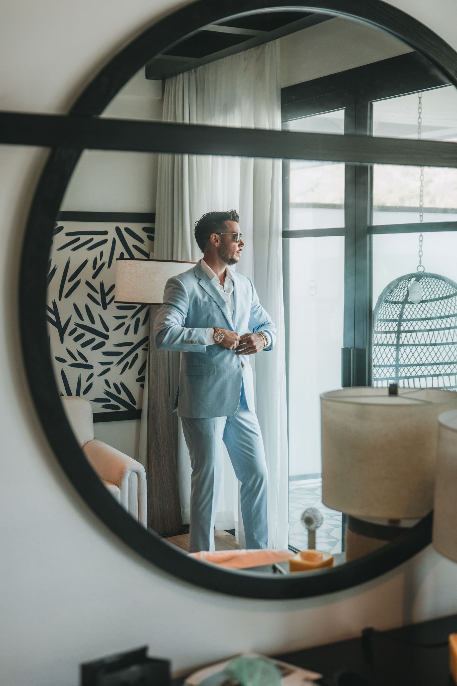 Man in light blue suit adjusting cufflinks reflected in round mirror in elegant hotel room.