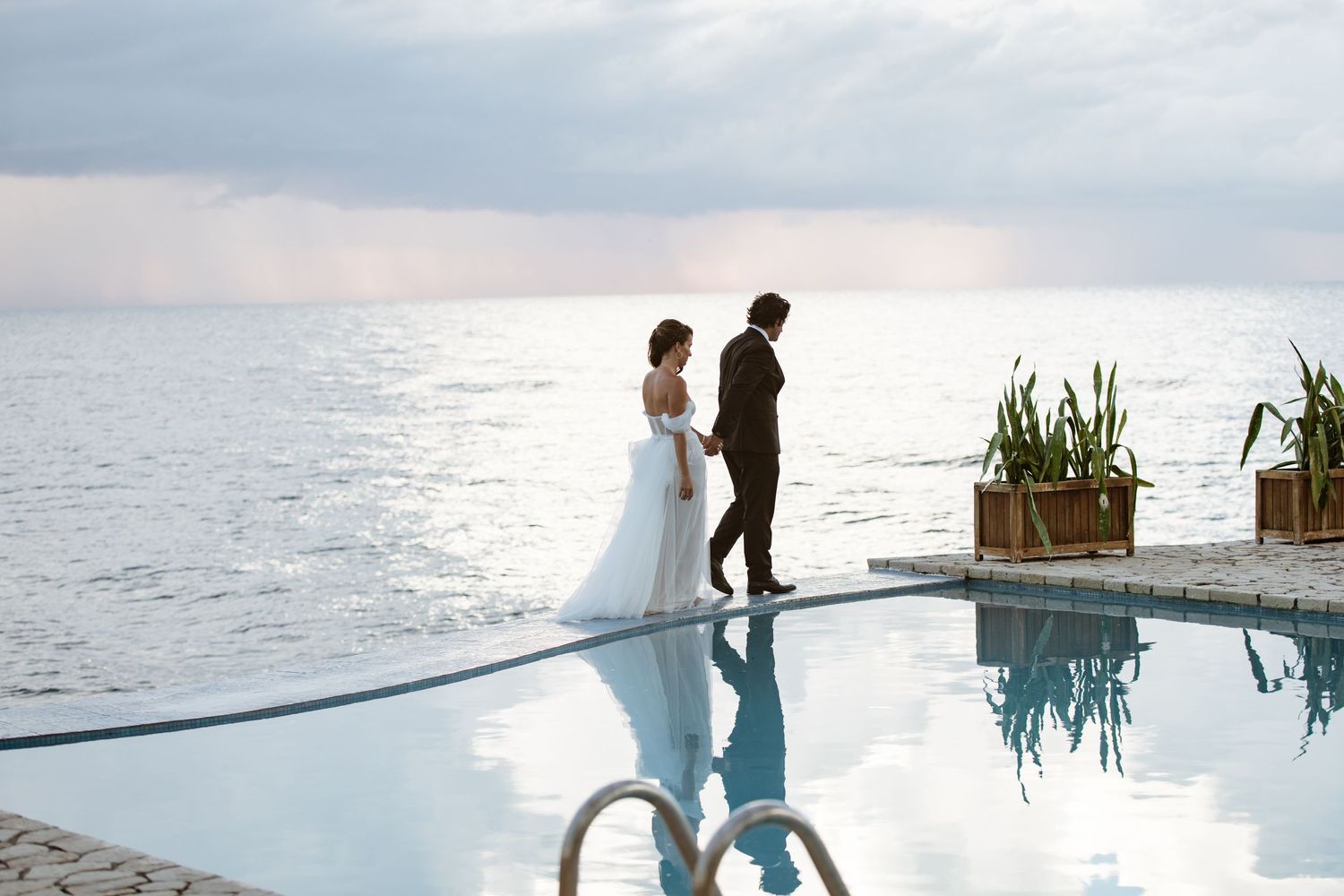 Bride and groom stand by infinity pool at sunset with dramatic ocean views on their wedding day.