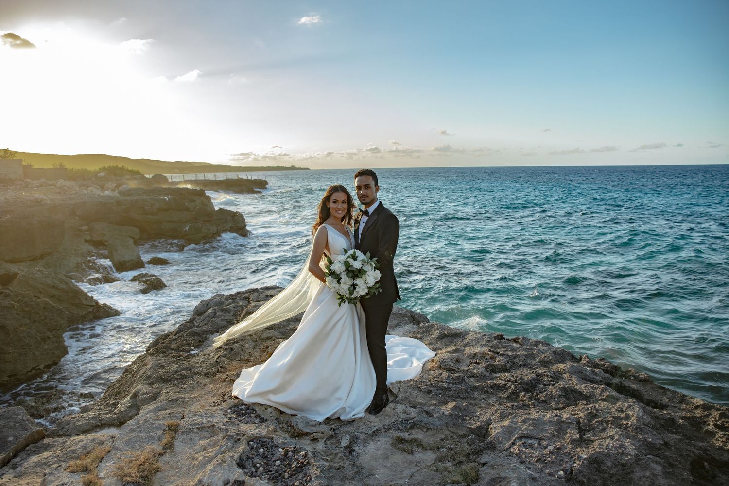 Newlyweds pose together on rocky coastal cliffside at golden sunset with turquoise ocean backdrop.
