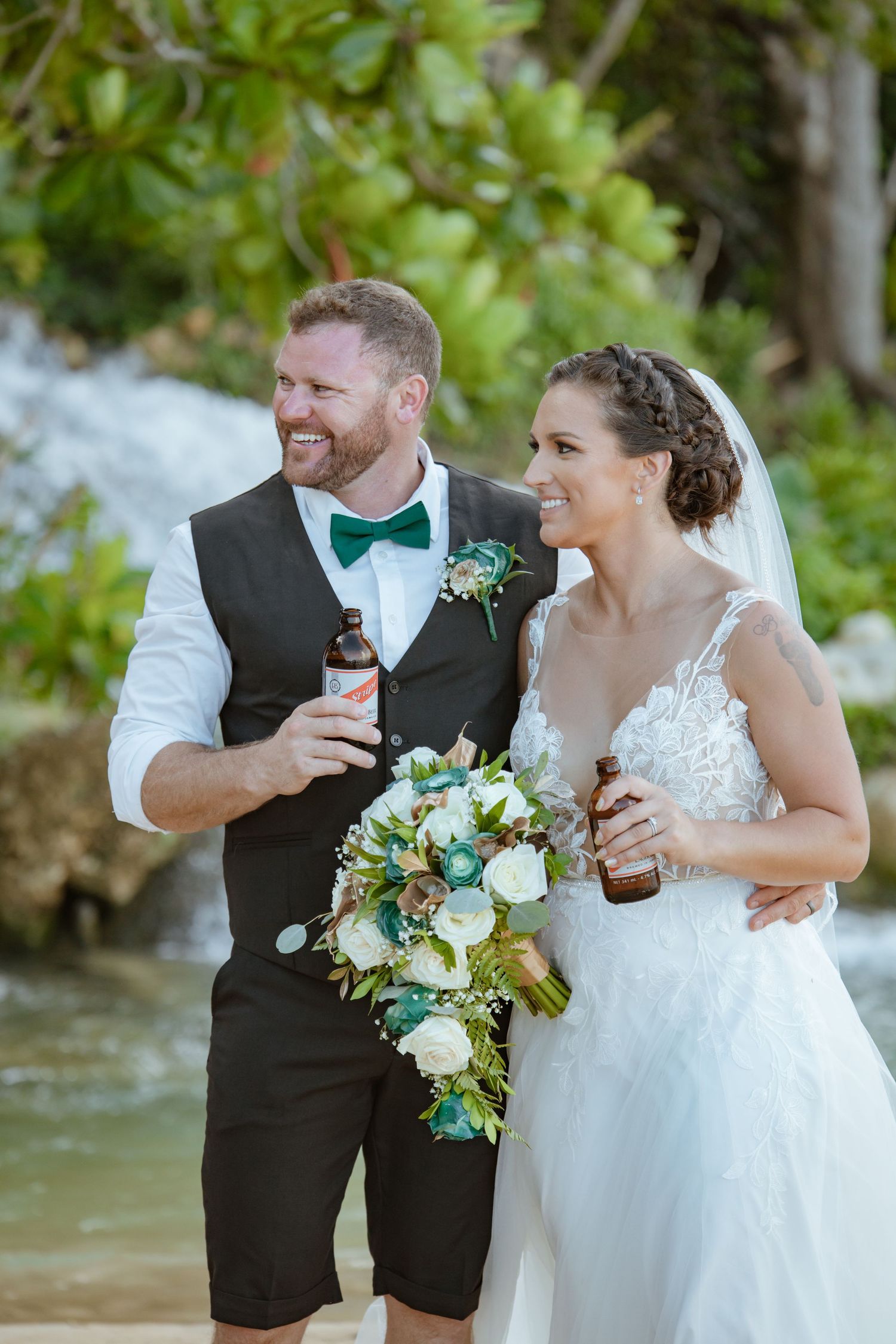 Smiling newlyweds holding beer bottles and a bouquet by a waterfall in a tropical setting.