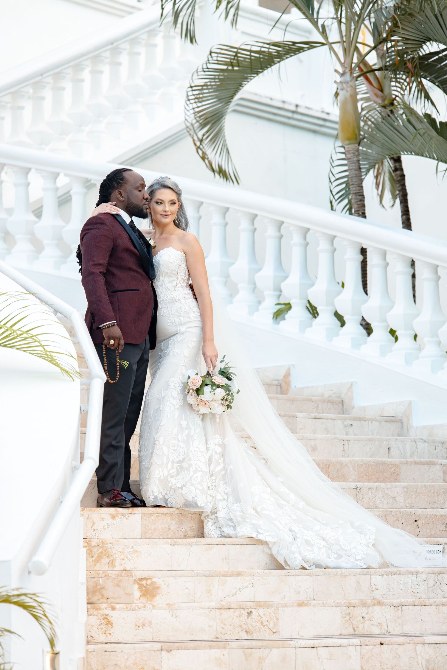Groom in burgundy suit kisses bride in lace mermaid gown on grand marble staircase.