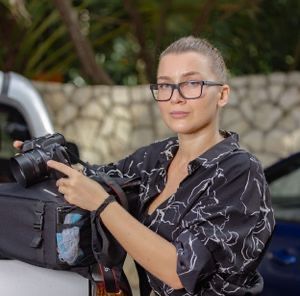 Female photographer with blonde hair and glasses holds professional camera outdoors at tropical location.