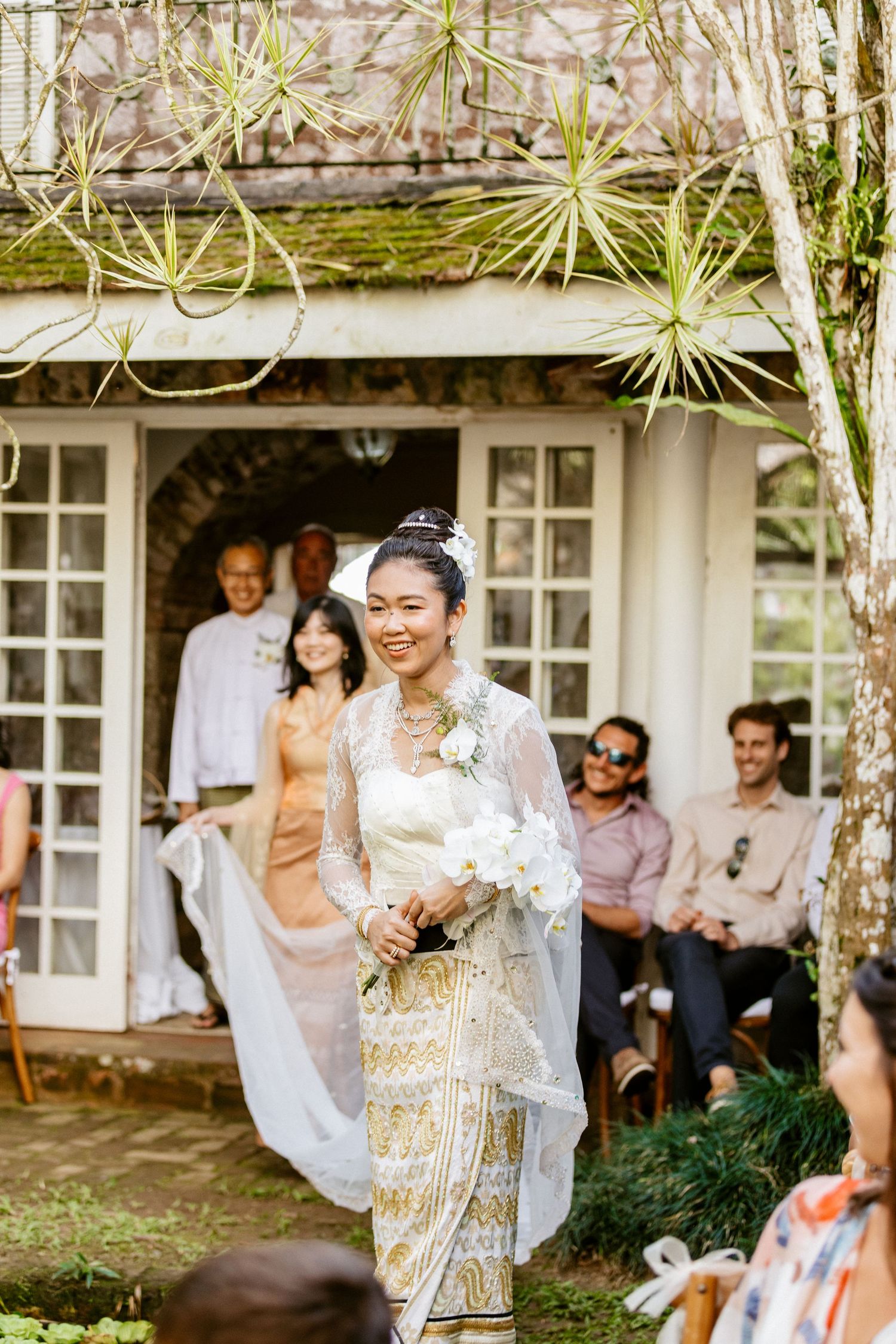 Smiling Asian bride in traditional gold and white dress walking down outdoor aisle with bouquet.