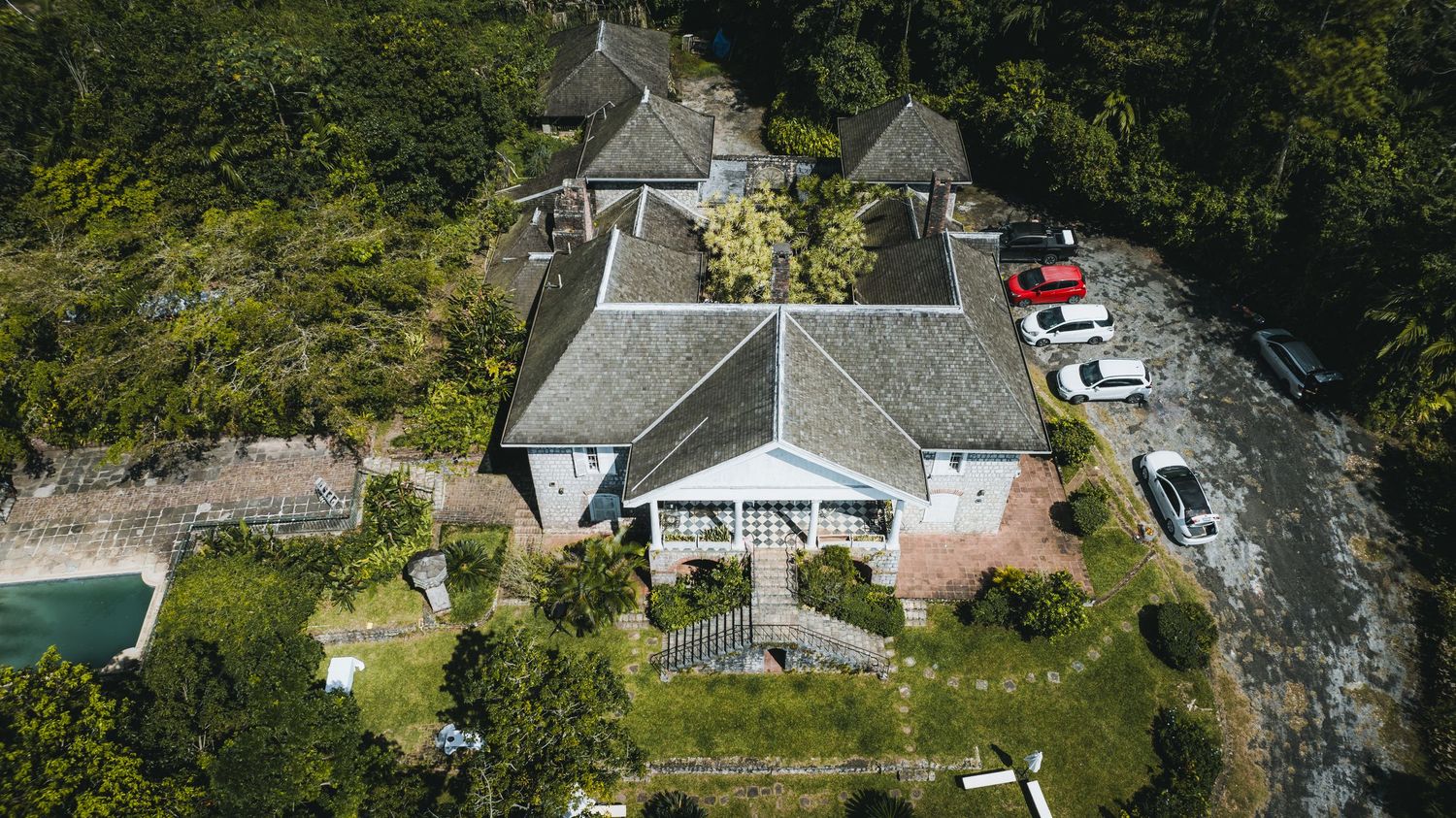 Aerial view of a large tropical villa with gray roofs surrounded by lush green trees and a parking area with cars.