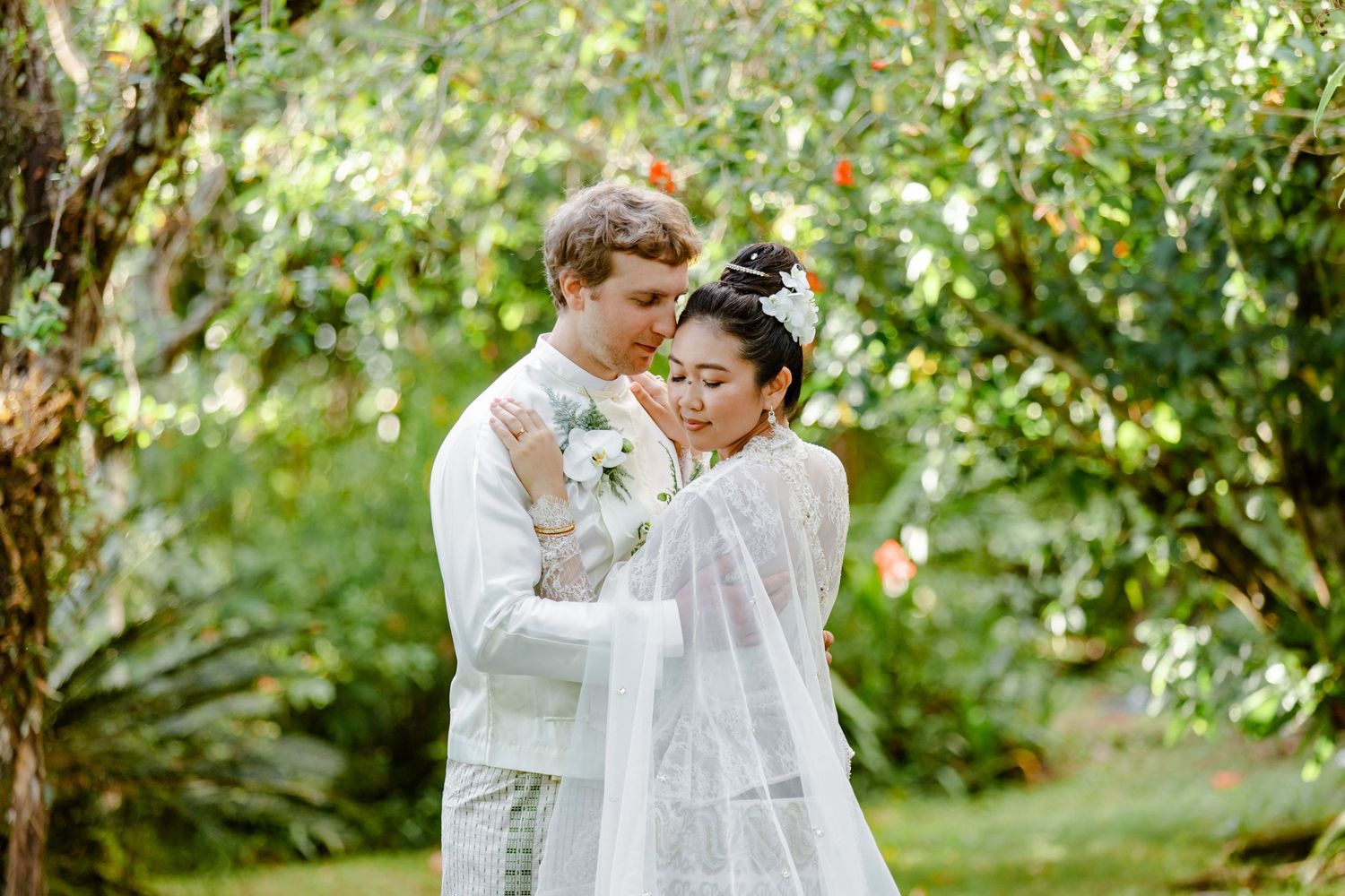 A couple in white wedding attire embraces tenderly in a lush green garden, the bride holding a bouquet.