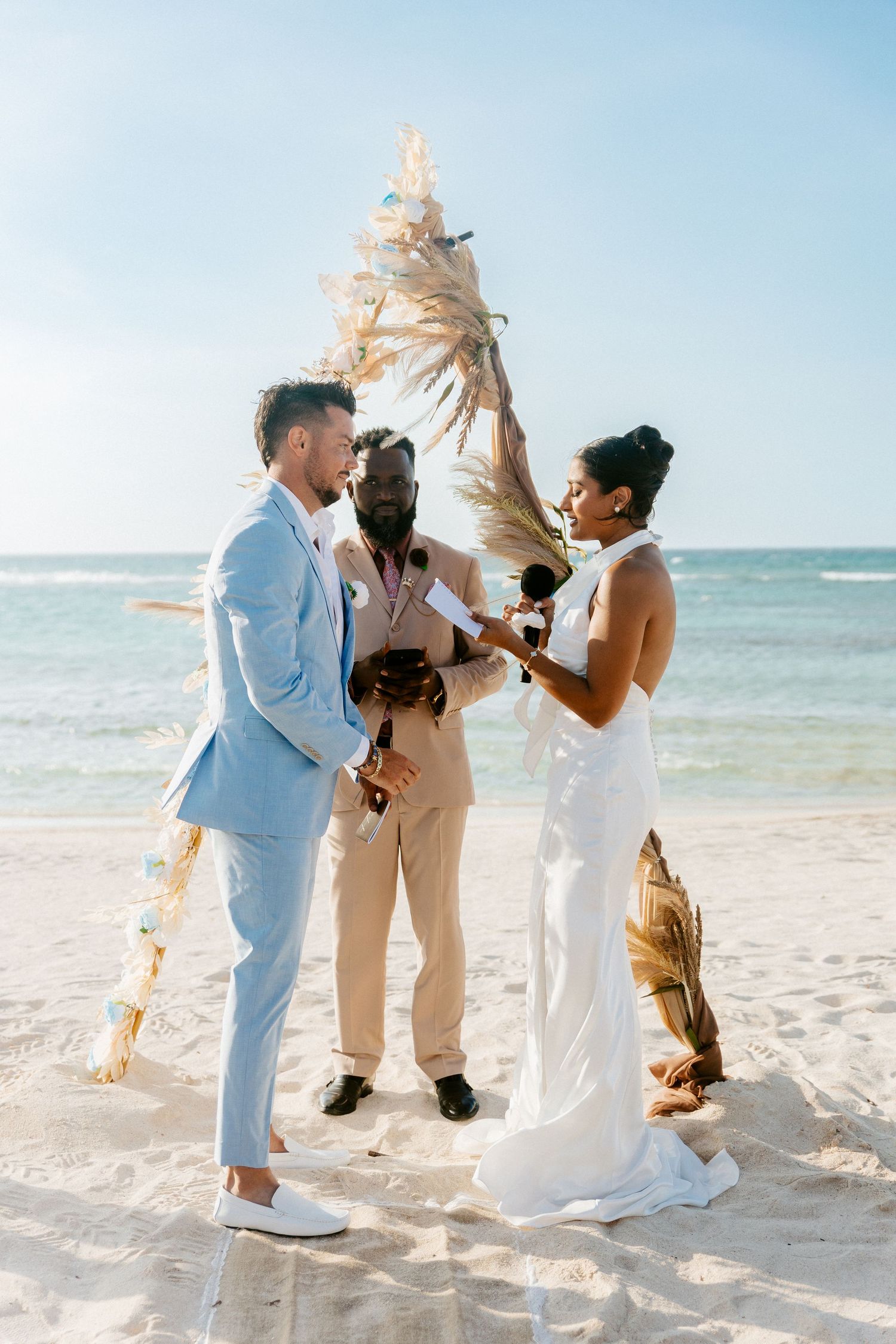 Bride in white halter gown reads vows to groom in light blue suit at tropical beach wedding ceremony.