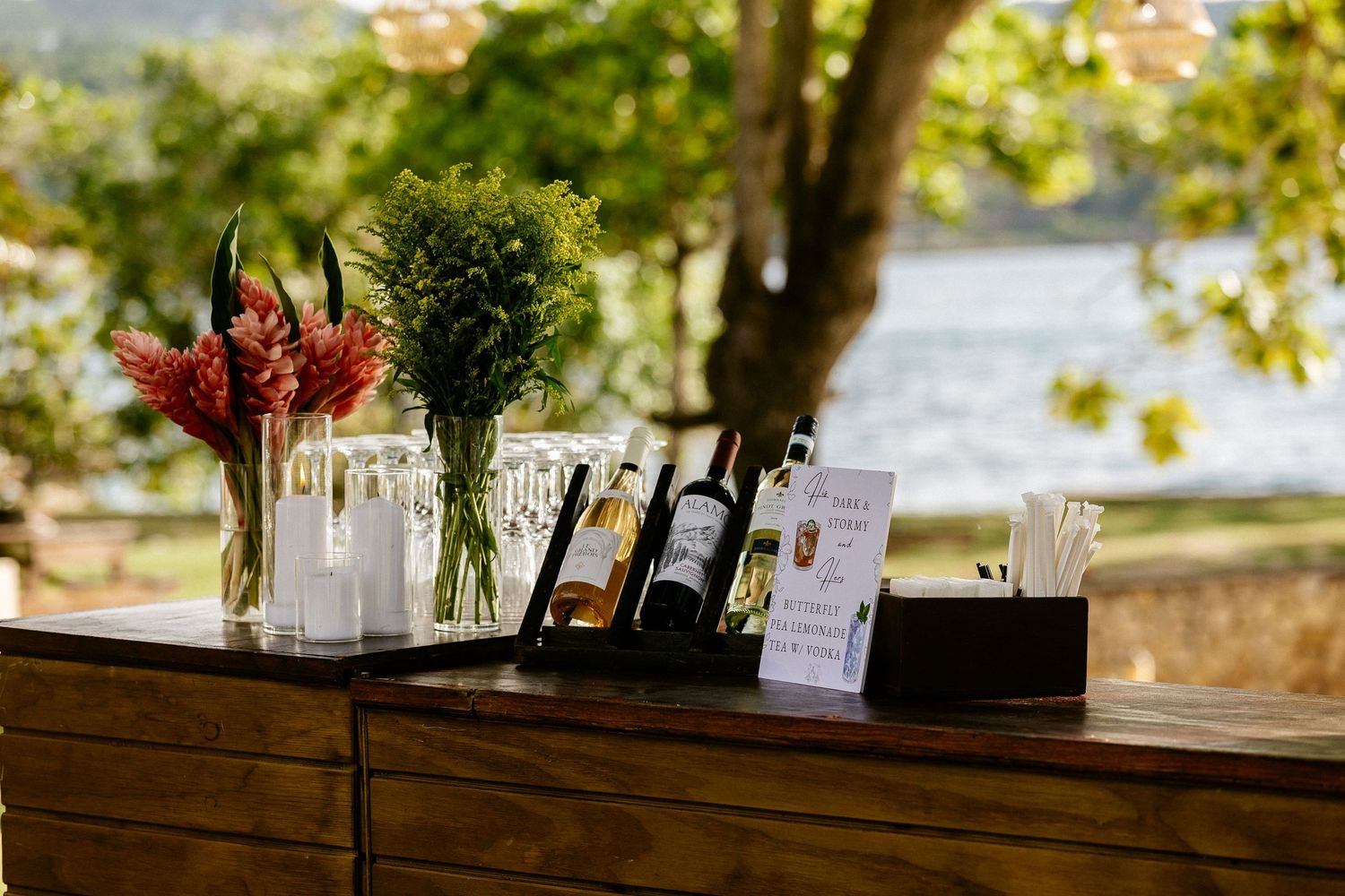 Outdoor bar setup with wine bottles, floral arrangements, and glassware on a wooden counter with a scenic waterfront backdrop.
