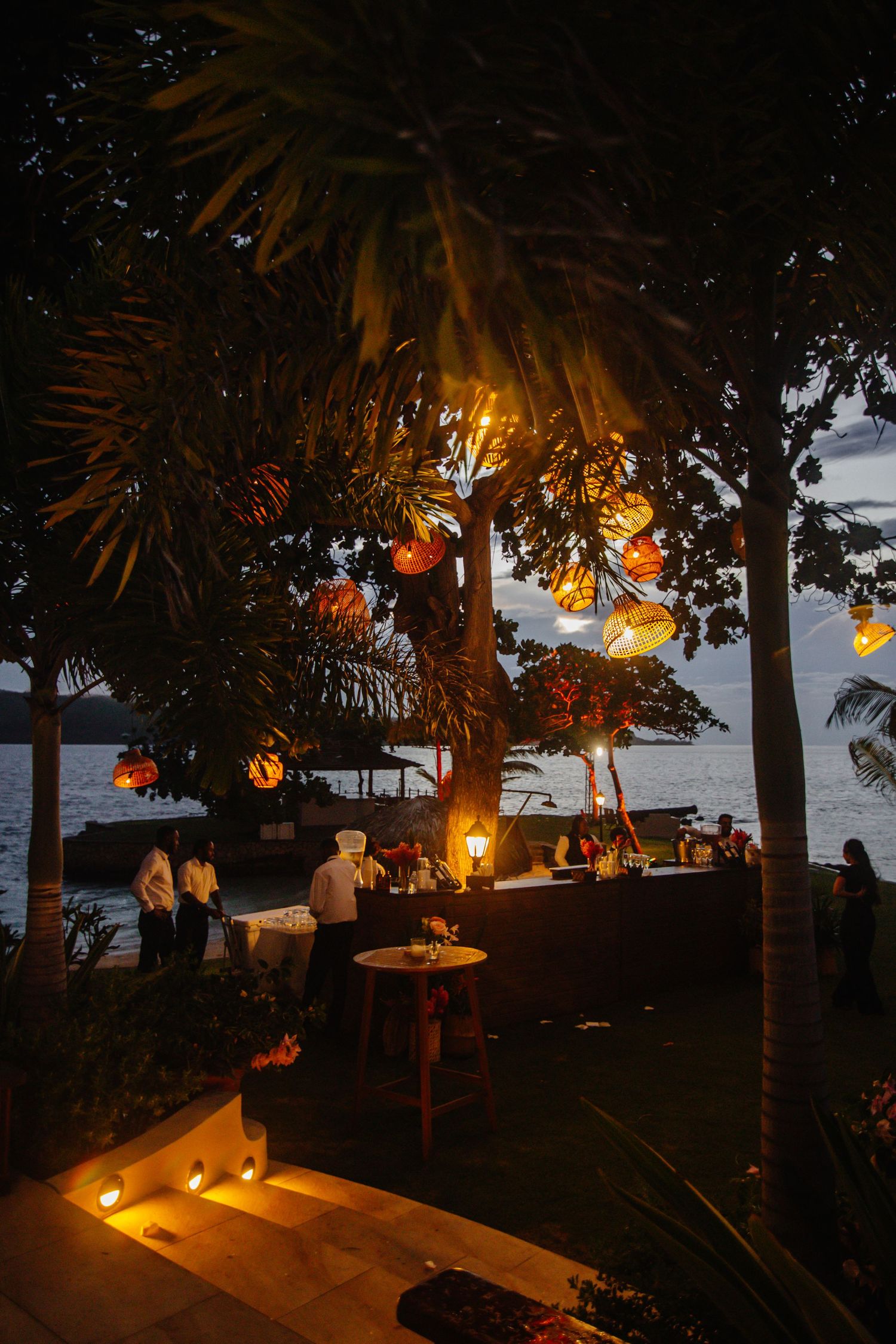 Tropical outdoor evening event with glowing lanterns hanging from palm trees near a waterfront venue at twilight.