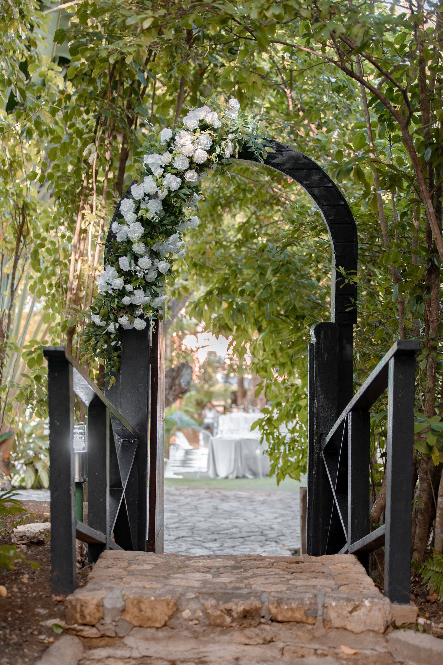 Black garden arch adorned with white roses and greenery framing an outdoor wedding venue entrance.