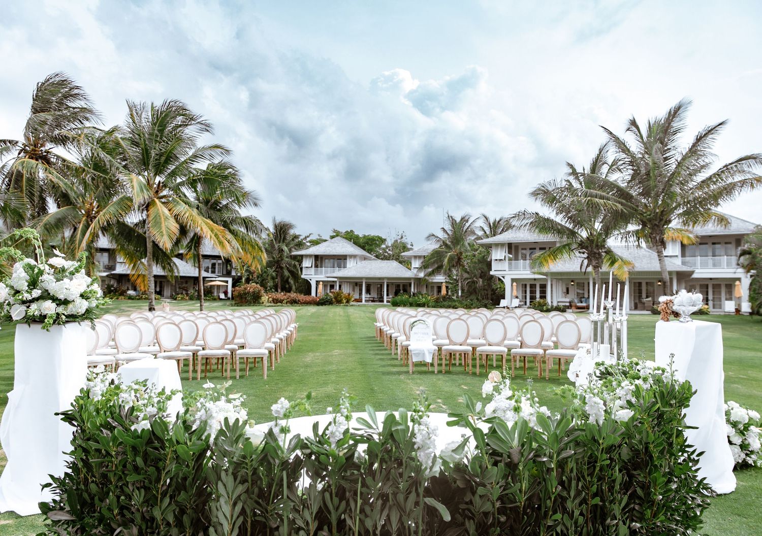 Elegant outdoor wedding ceremony setup with white chairs, floral arrangements, and palm trees at a tropical resort.