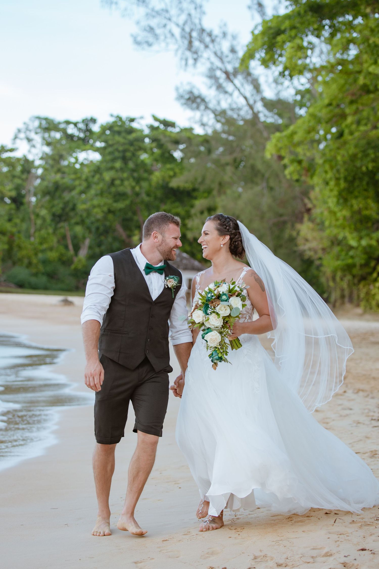 Bride and groom walking hand in hand on a beach, veil flowing in the breeze.