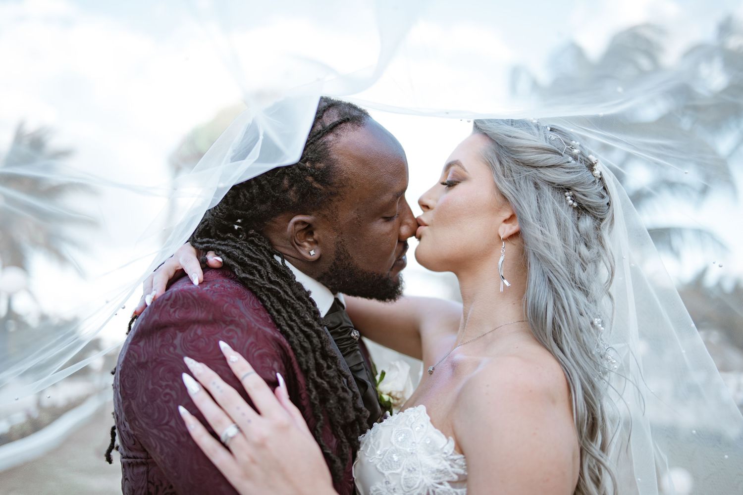 Interracial newlyweds share a romantic kiss under a flowing white veil at their outdoor wedding.