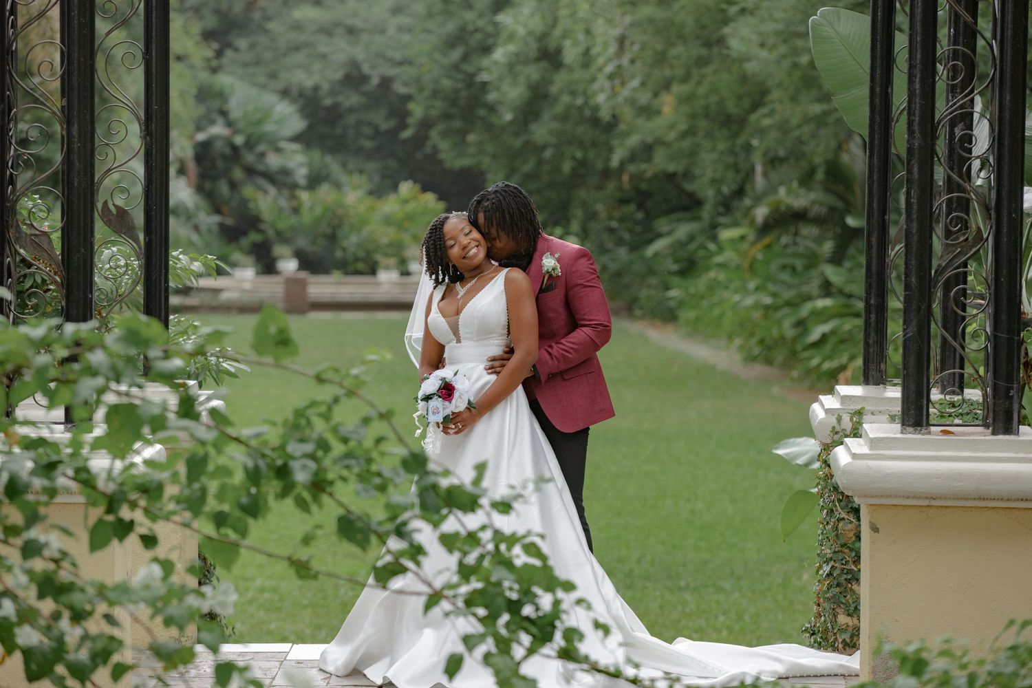 Black couple shares romantic wedding portrait in lush garden gazebo, groom in burgundy suit embracing bride in white gown.