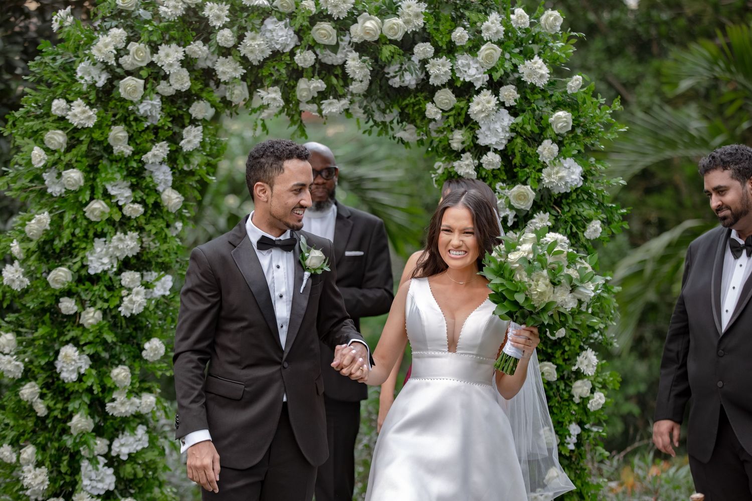 Smiling newlyweds walk under floral arch of white flowers and greenery after wedding ceremony.