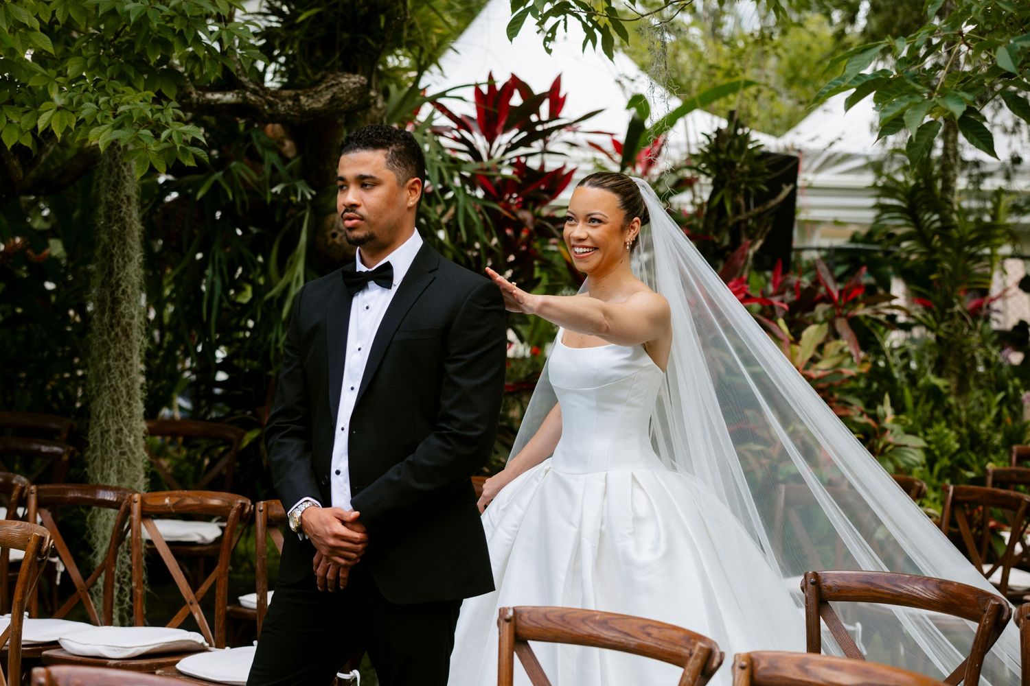 A bride in a strapless white gown and veil touches her groom's shoulder during an outdoor garden wedding.