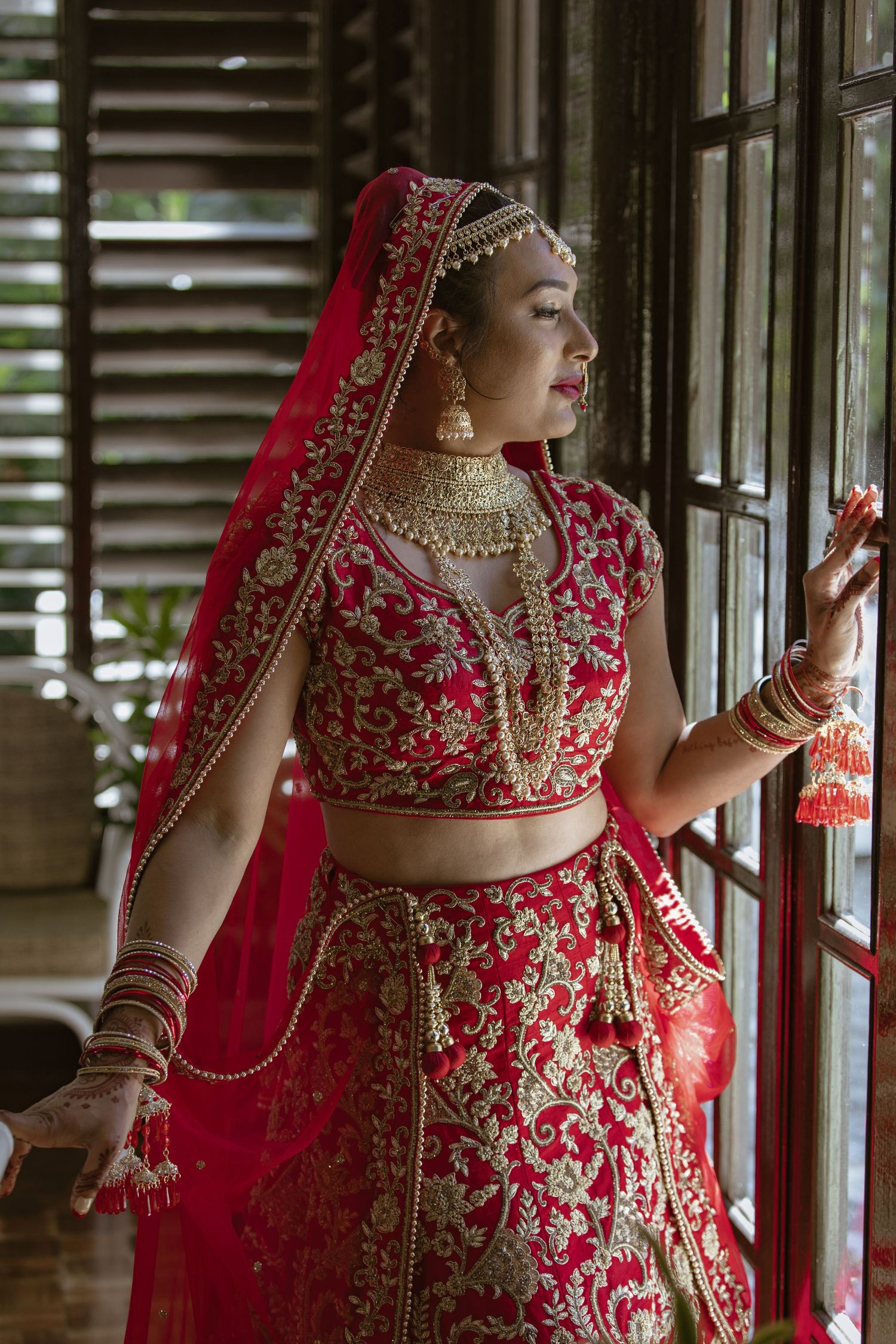 Indian bride in ornate red and gold lehenga with traditional jewelry gazes through wooden window shutters.