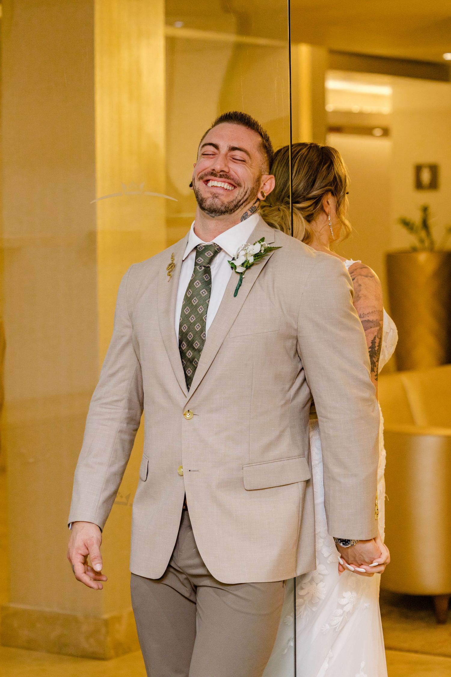 Smiling groom in beige suit with green tie and boutonniere stands with bride in lace wedding dress during ceremony.