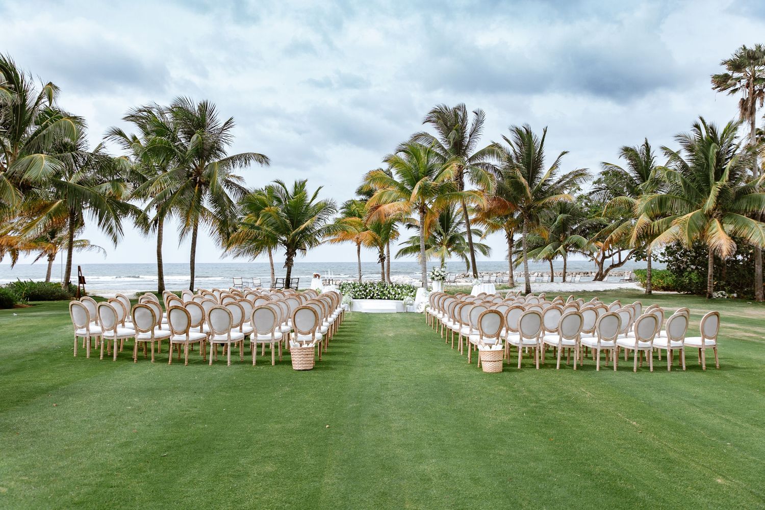 Elegant outdoor wedding ceremony setup with gold chairs on lush green lawn surrounded by tropical palm trees near the ocean.
