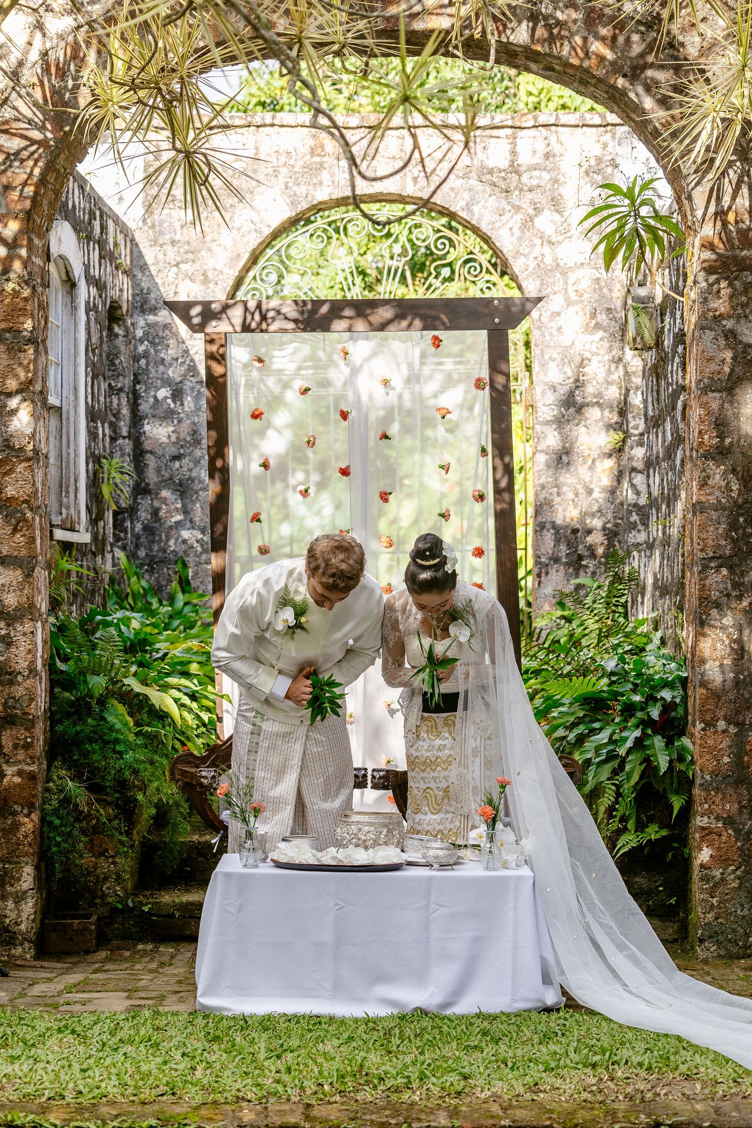 Bride and groom cutting wedding cake under stone arch with lush greenery and floral backdrop outdoors.