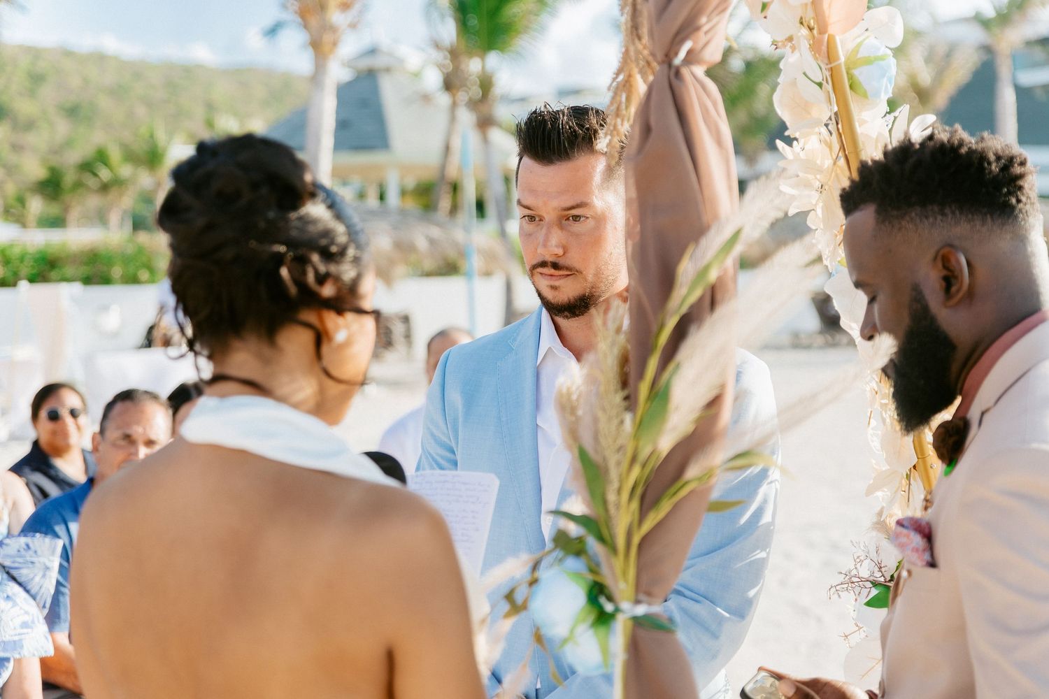 Groom in light blue suit exchanges vows at outdoor beach wedding ceremony with officiant and guests.