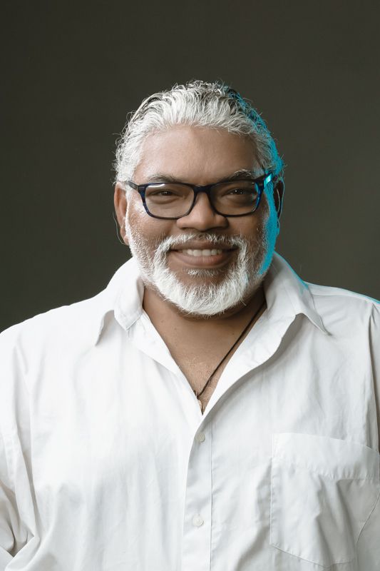 A smiling older man with white hair and beard wearing glasses and a white shirt poses for portrait.