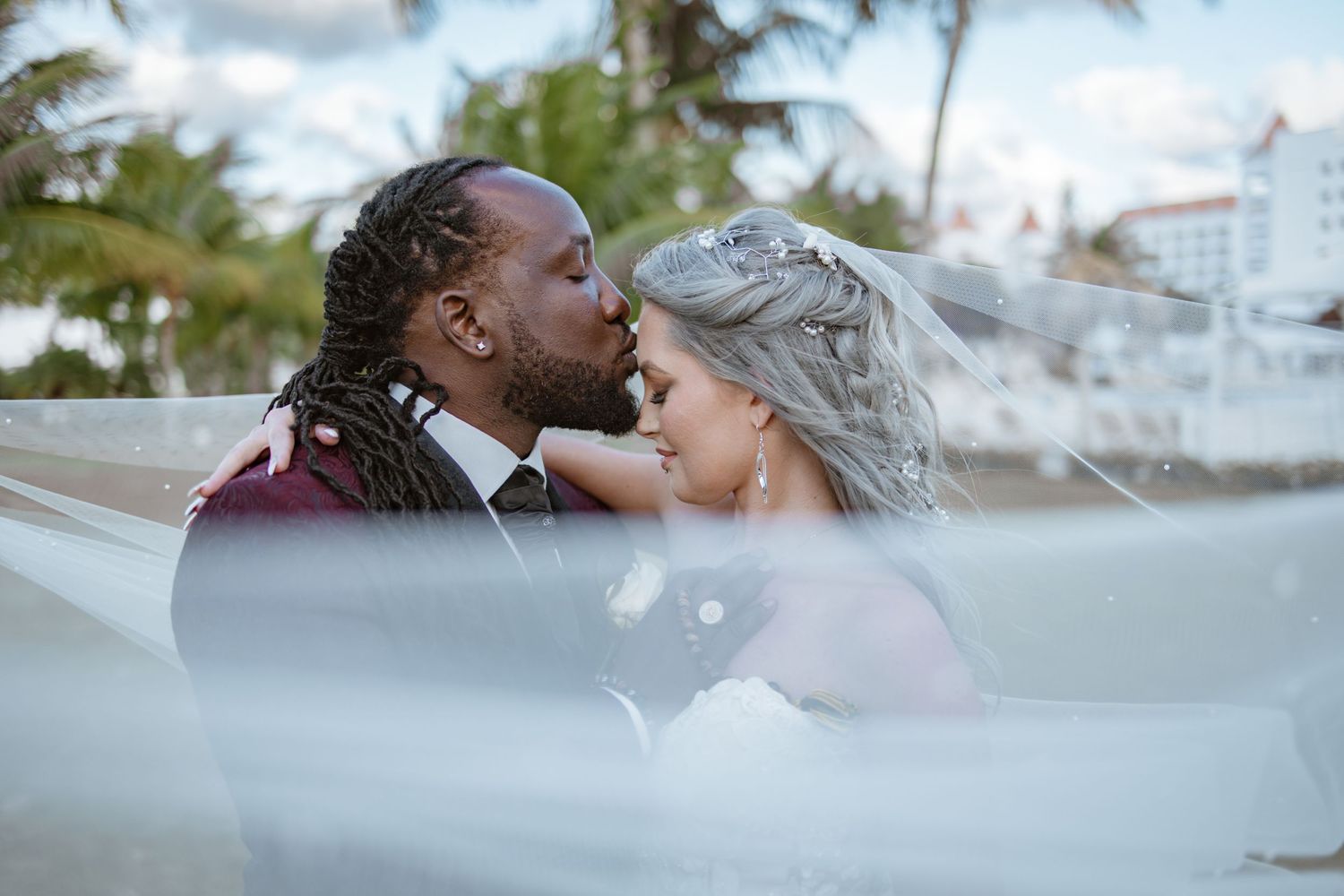 Newlywed couple shares a kiss on the beach framed by a billowing white wedding veil.