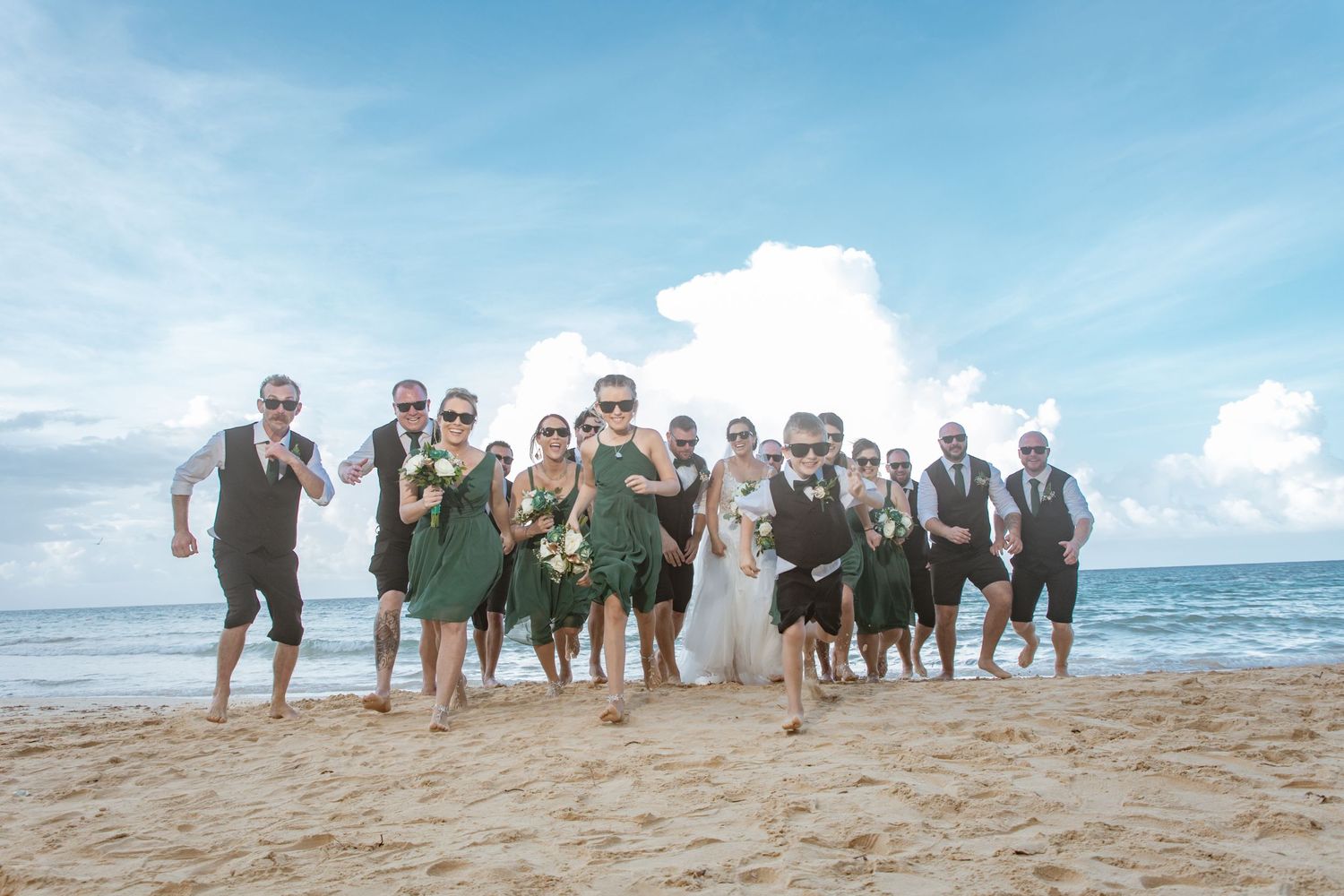 Joyful wedding party running and posing together on a sunny tropical beach shoreline.